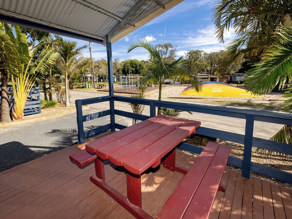 A wooden picnic table with benches under a covered area, surrounded by palm trees and a sandy area with a yellow object. - BIG4 Manning Point Holiday Park (Manning Point)