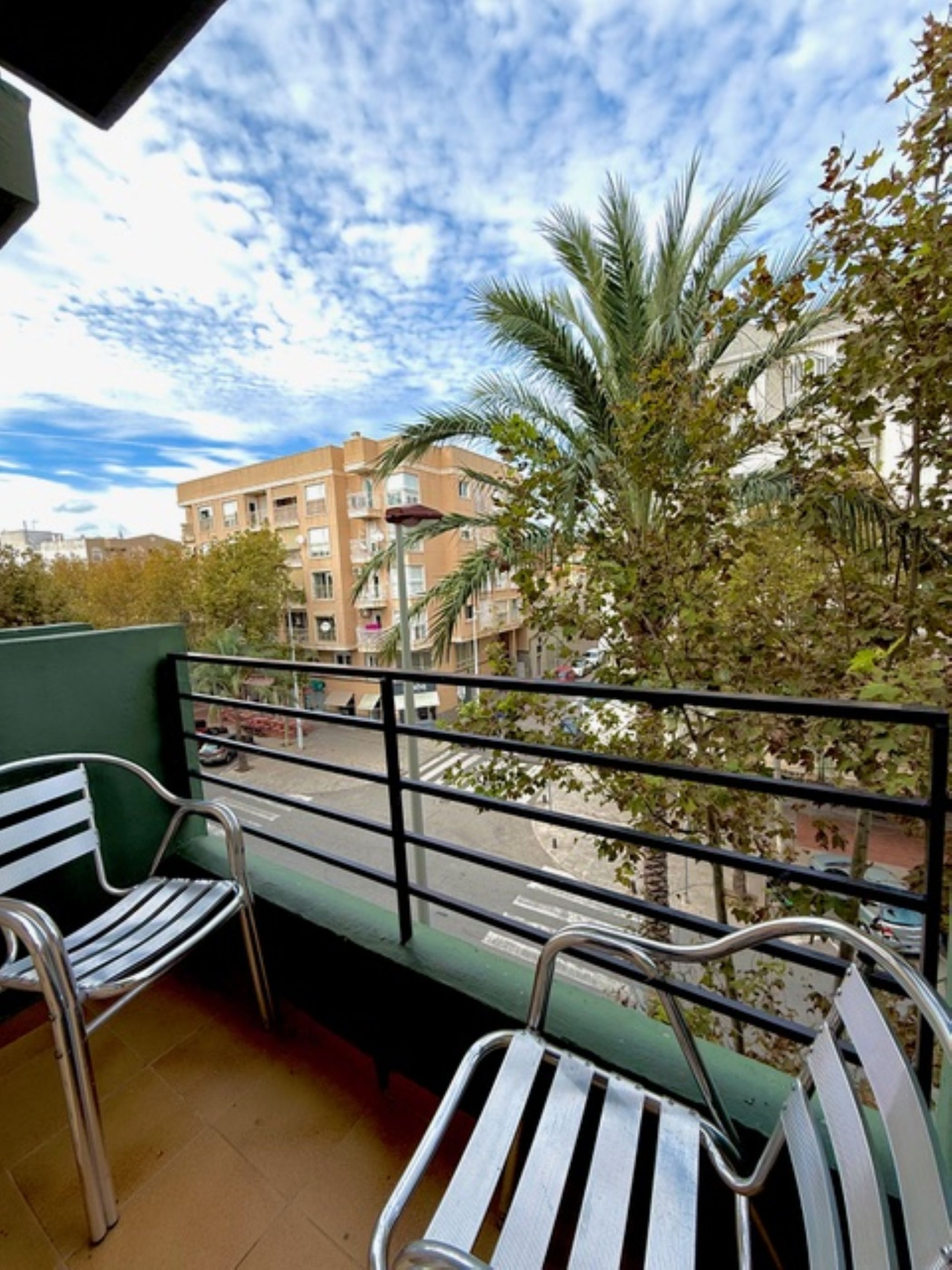 A balcony with metal chairs, a view of a building, and a palm tree.