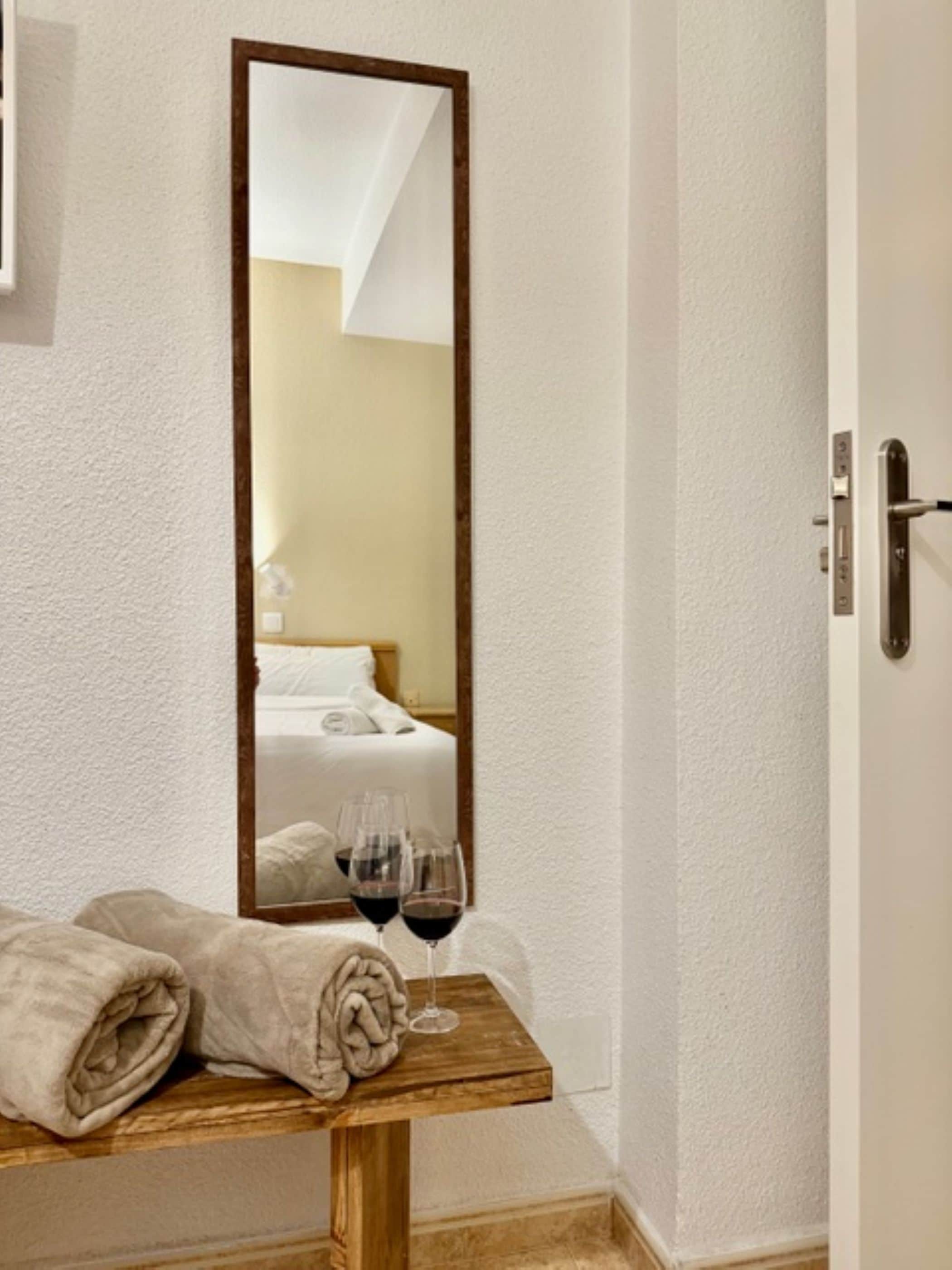 A hotel room with a wooden bedside table, a mirror, a bed with white linens, and a glass of red wine.
