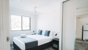 A modern bedroom with a large bed, a nightstand, a bedside table, a ceiling fan, and a window with a view of trees. - BIG4 Rainbow Beach Holiday Park (Rainbow Beach)