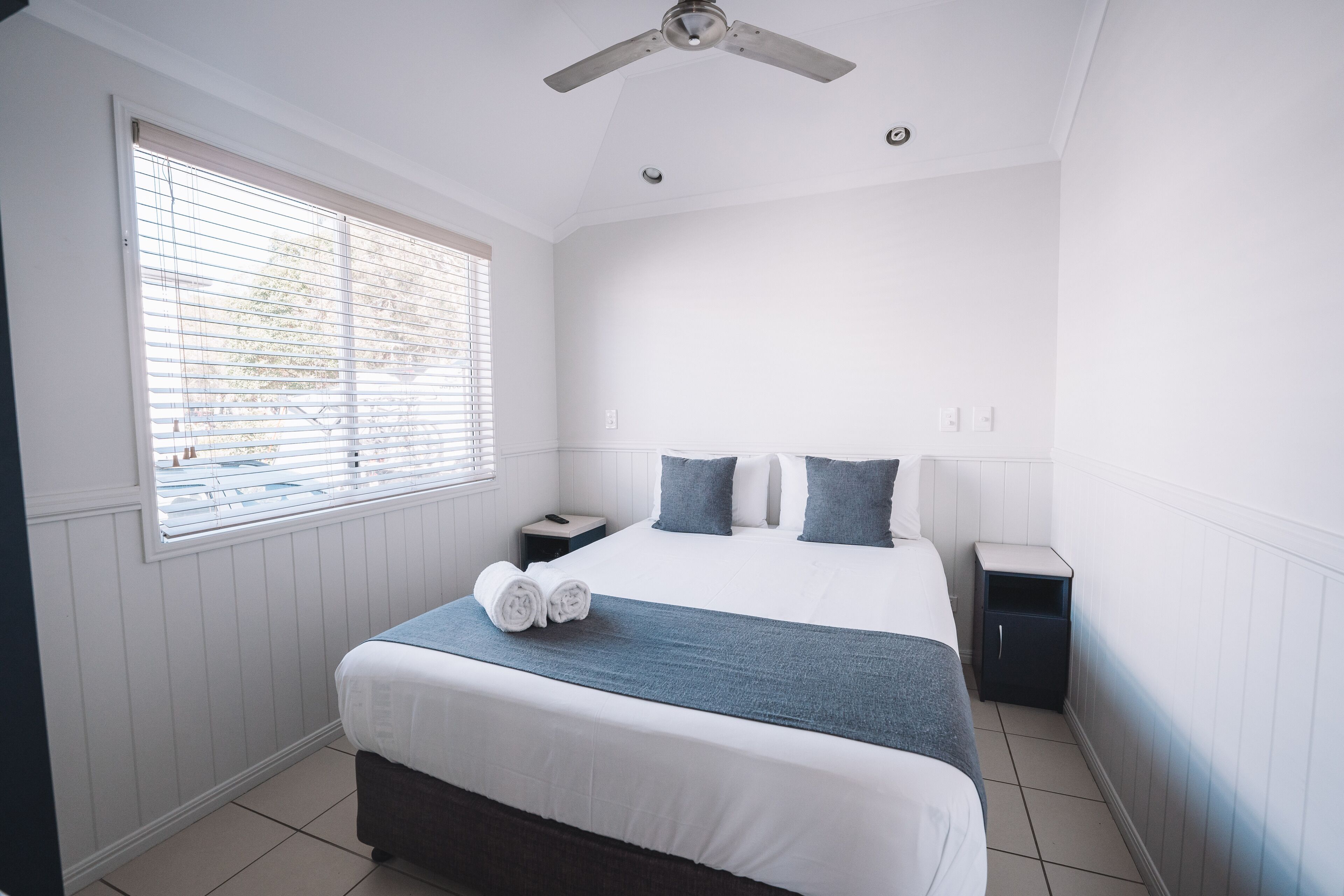 A bedroom with a large bed, two bedside tables, a ceiling fan, and a window with blinds.