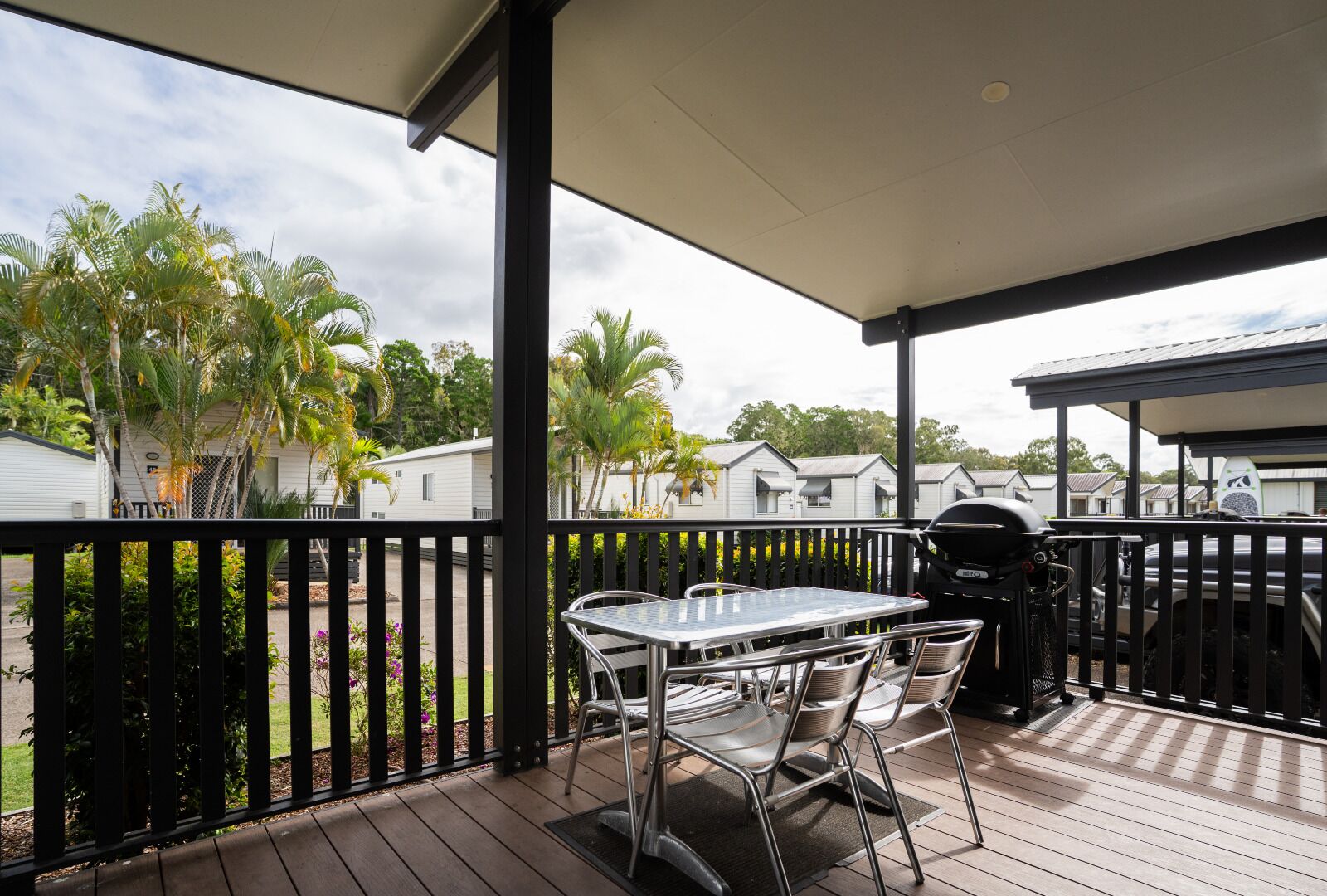 A covered outdoor area with a table and chairs, a grill, and a view of mobile homes and palm trees.