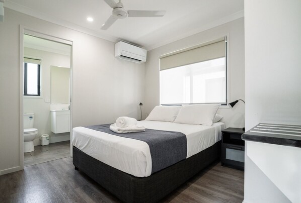 Una habitación de hotel con cama, ventilador de techo, aire acondicionado, ventana con persianas, lámpara de noche y una mesita.