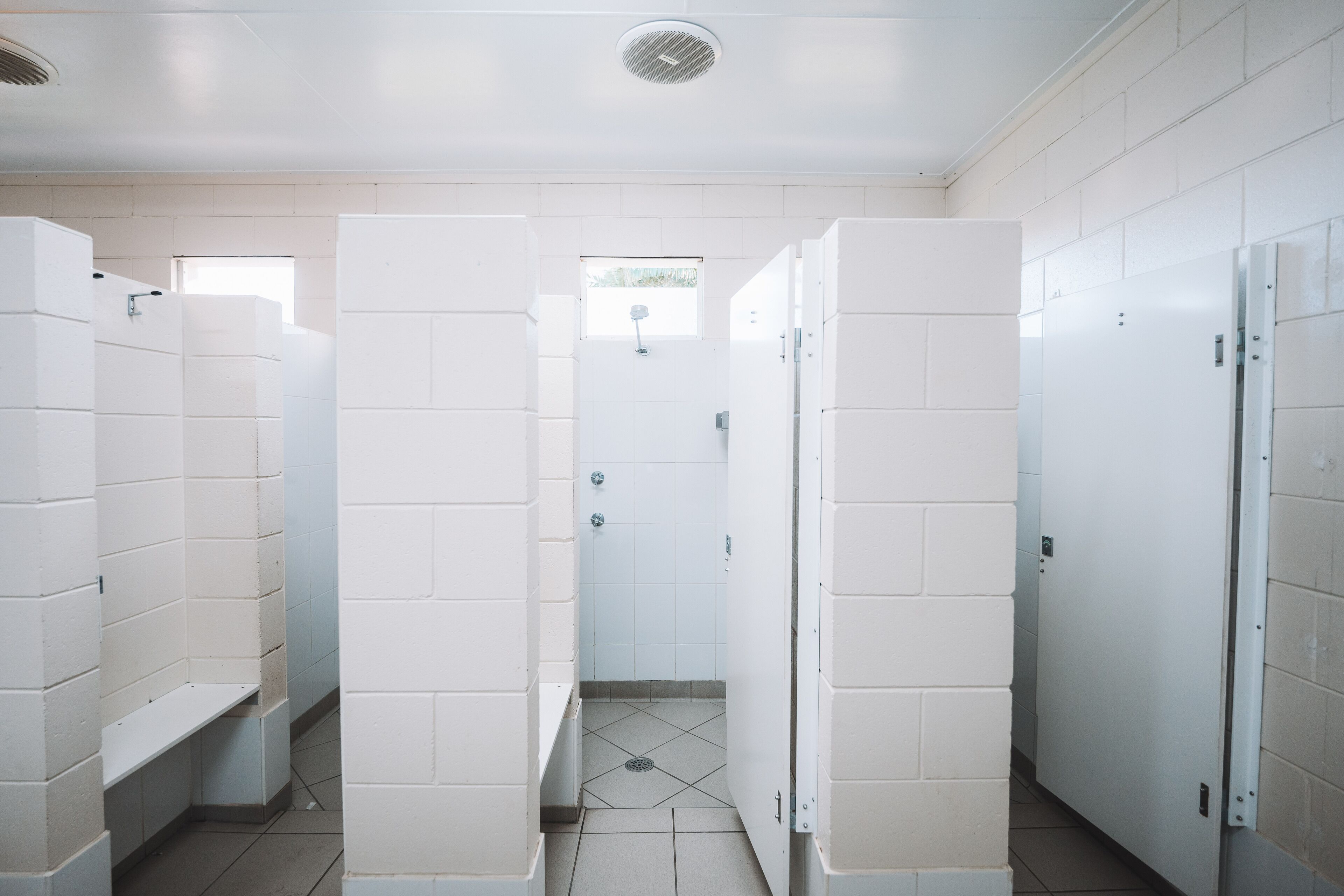 A public restroom with white tiled walls and floors, multiple stalls, and a visible shower area.