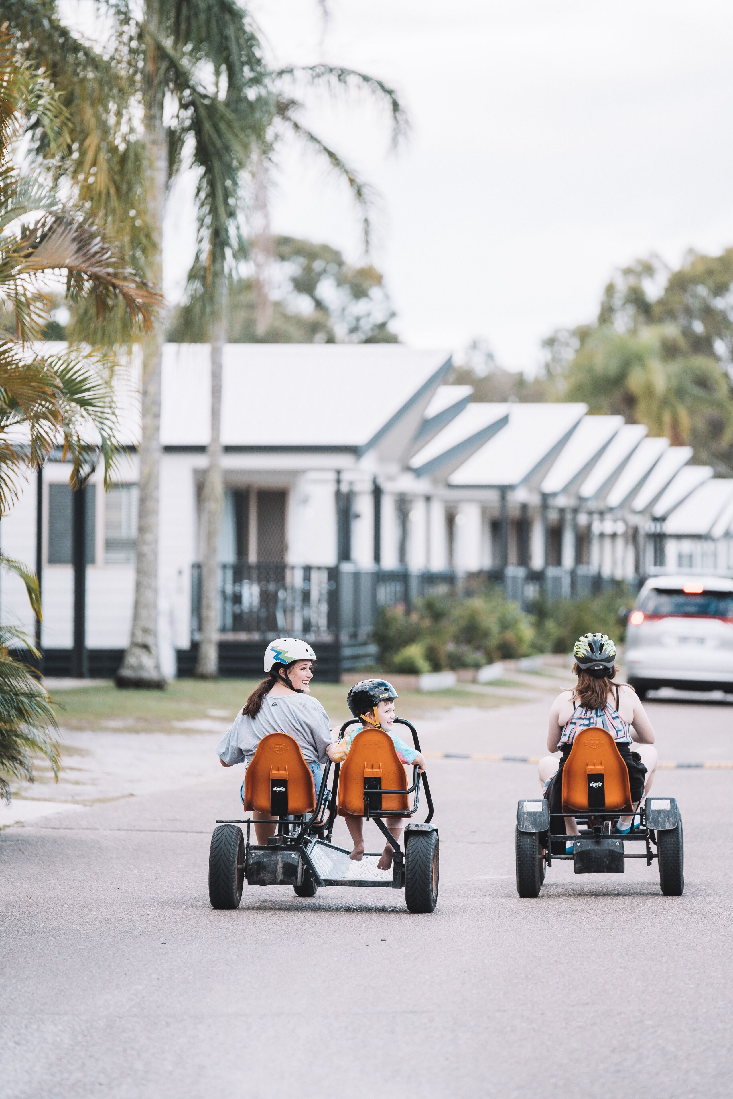 Three people in go-karts on a street with houses in the background.