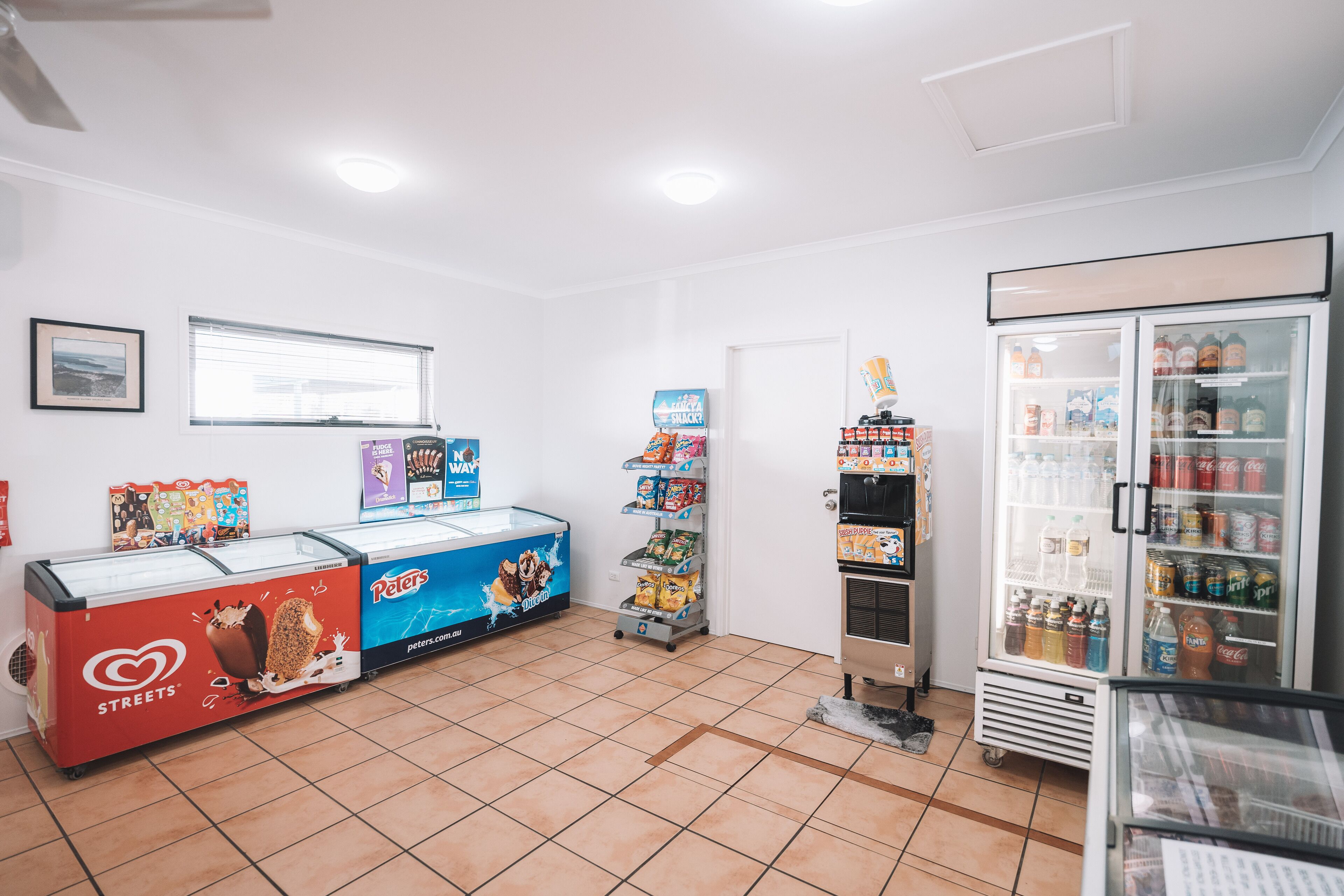 A clean, well-lit convenience store with a freezer section, snack shelves, and a refrigerated beverage display.