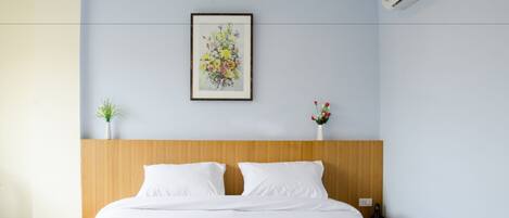 Habitación de hotel con cabecera de madera, cama con ropa de cama blanca, un cuadro con motivos florales en la pared, una pequeña planta en maceta y un jarrón con flores rojas.