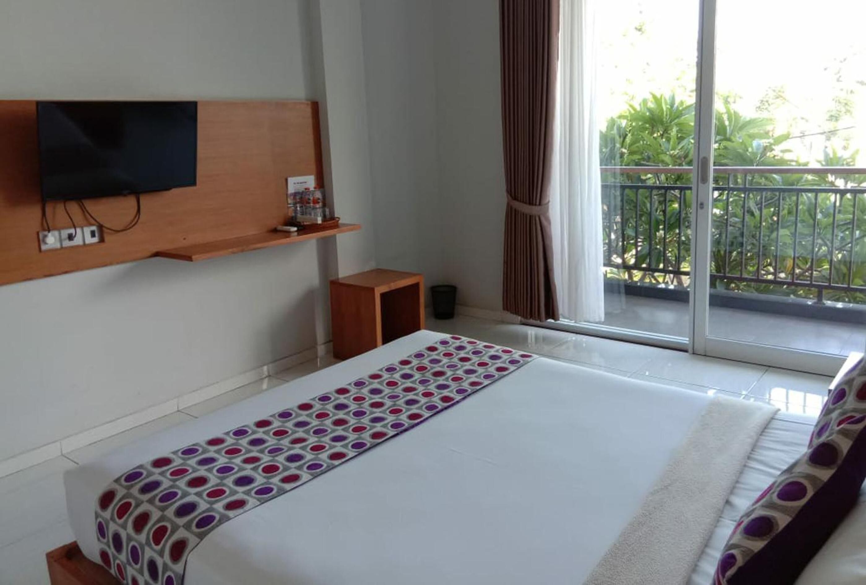 Een hotelkamer met een groot bed, een aan de muur bevestigde televisie, een klein houten tafeltje en een balkon met uitzicht op groen.