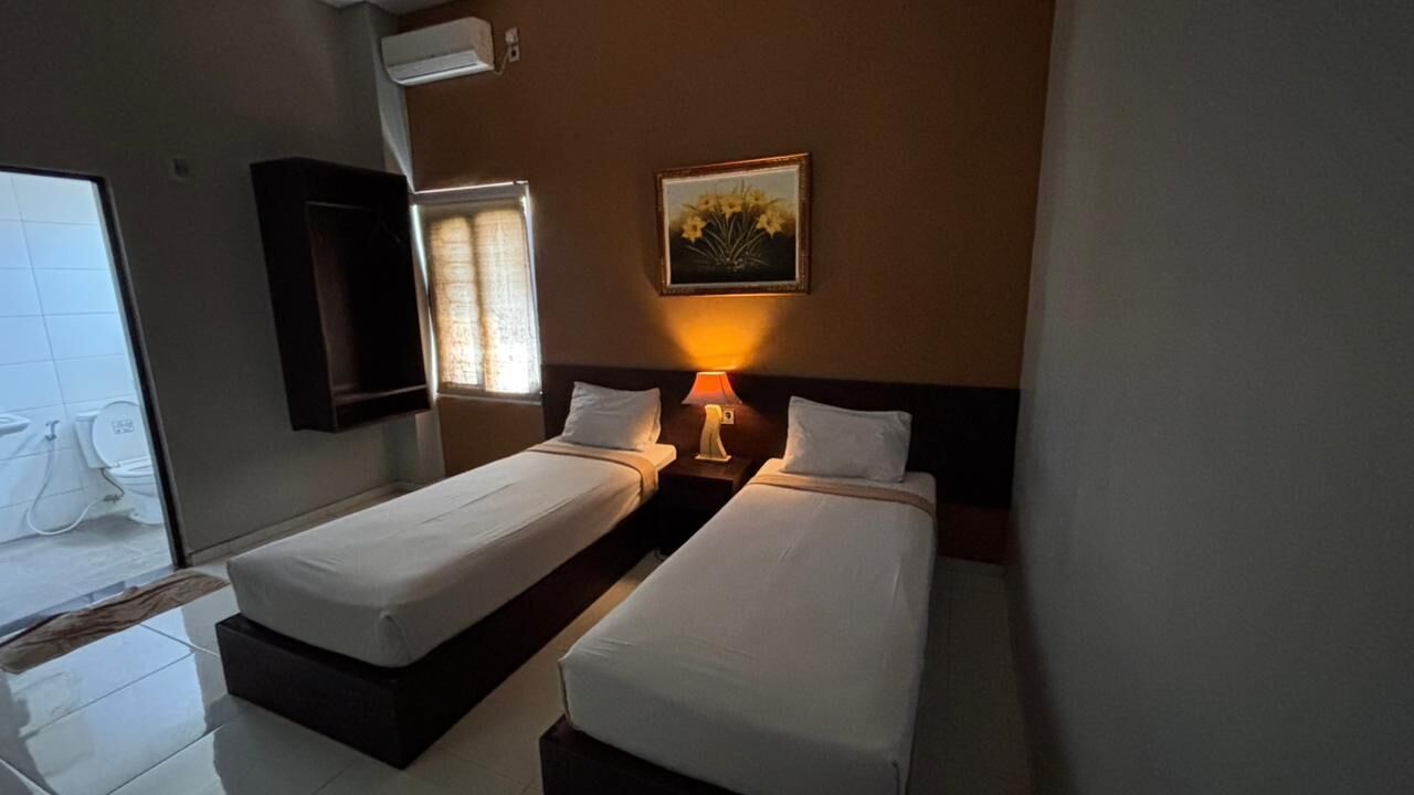 Ein Hotelzimmer mit zwei Betten, einem Nachttisch mit Lampe, einem Bild an der Wand und einem Fenster mit Vorhängen.