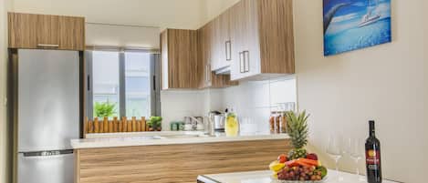 A modern kitchen with wooden cabinets, a stainless steel refrigerator, a dining table with a fruit bowl and wine glasses, and a painting of a boat on the wall.