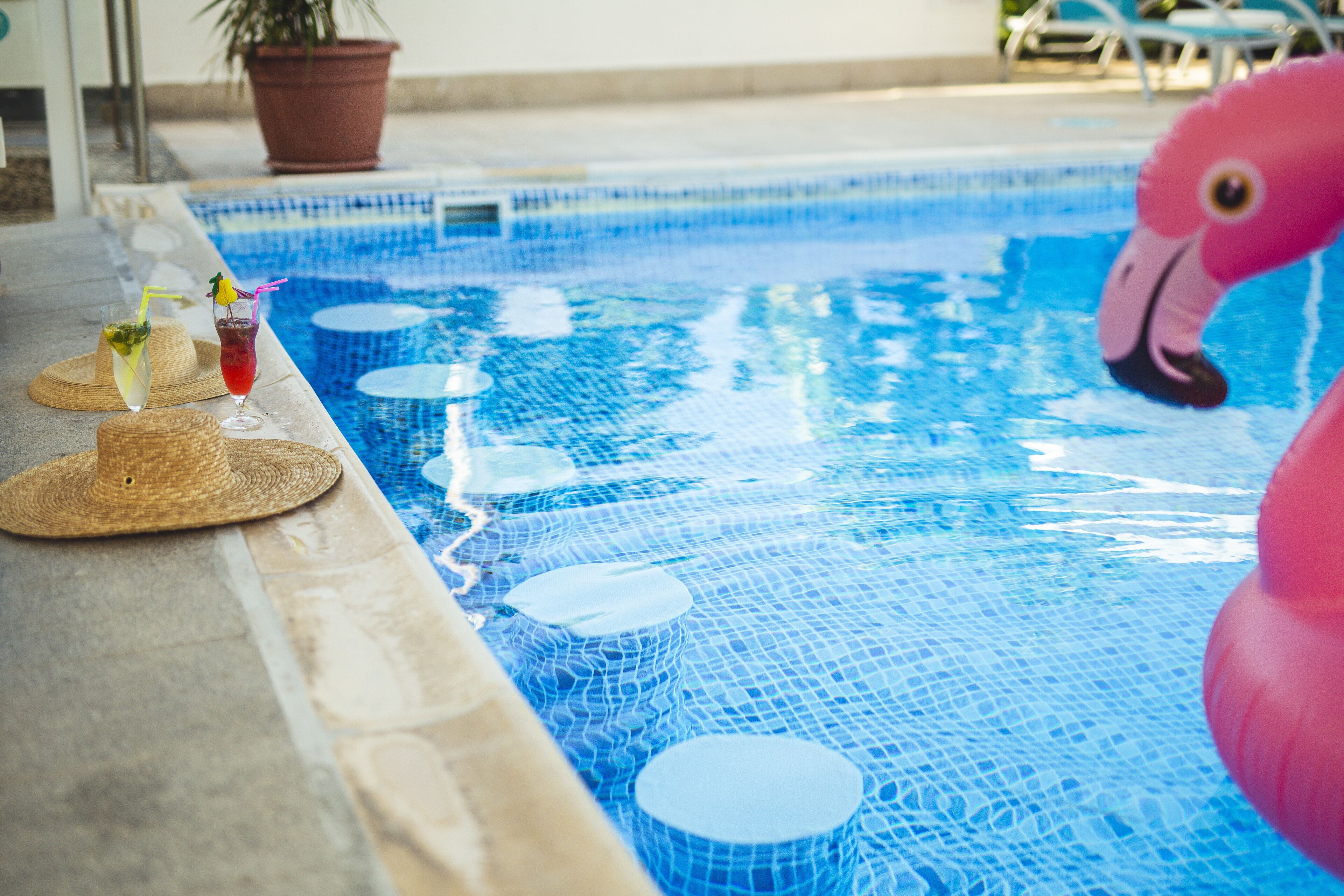 A swimming pool with a pink flamingo float, a hat, and a drink on the edge.