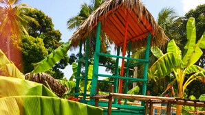 A playground with a thatched roof structure, surrounded by banana plants and palm trees. - Saara Hotel (Mahajanga)