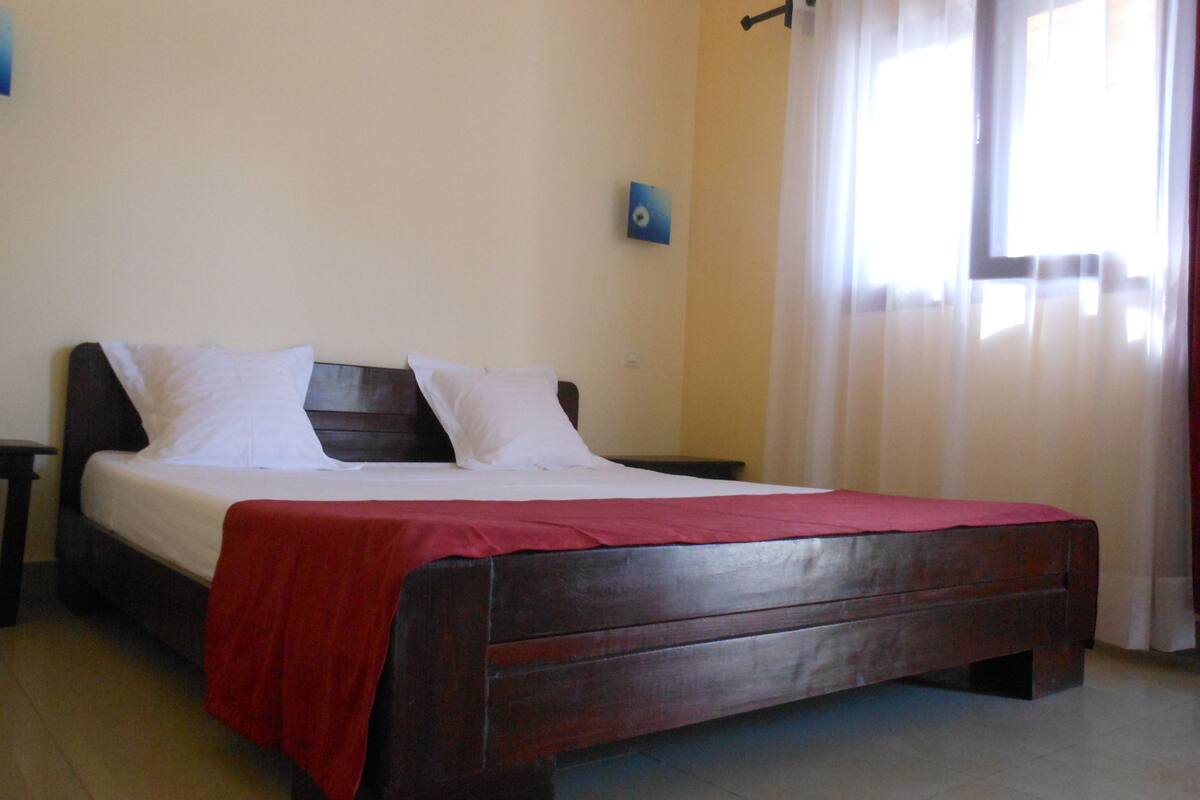Ein Bett mit hölzernem Gestell, weißer Bettwäsche, einer roten Decke und einem kleinen Nachttisch.
