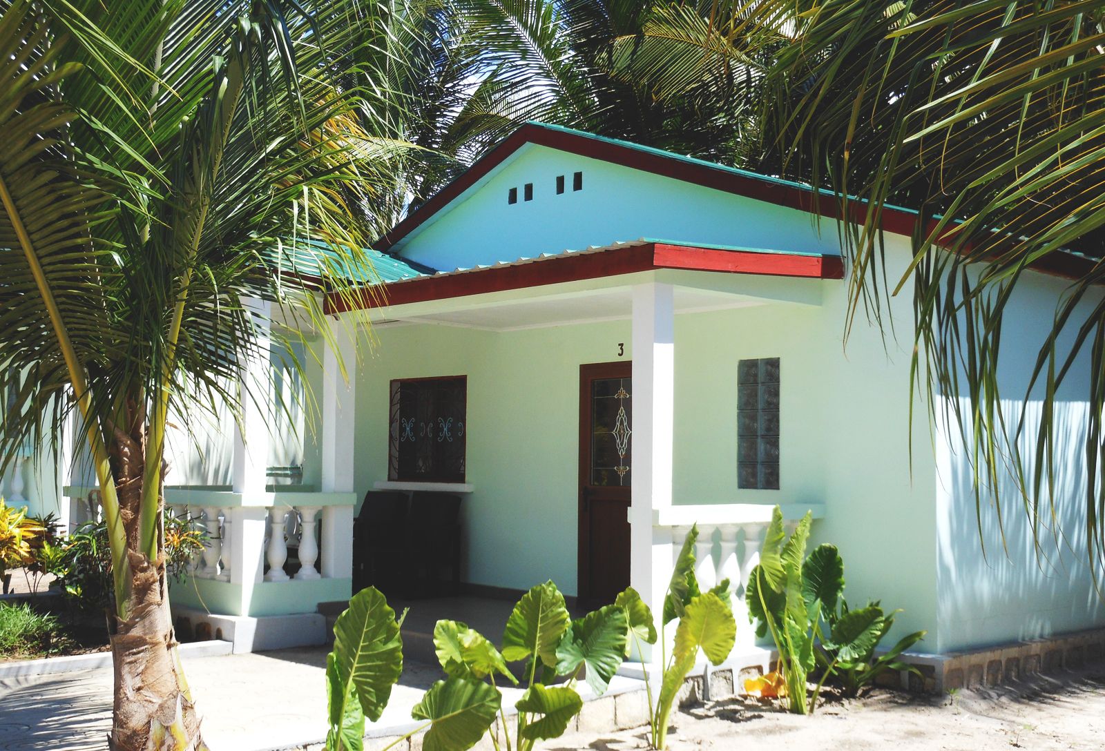 Una casa de un piso con fachada azul claro y techo rojo, rodeada de abundante vegetación.