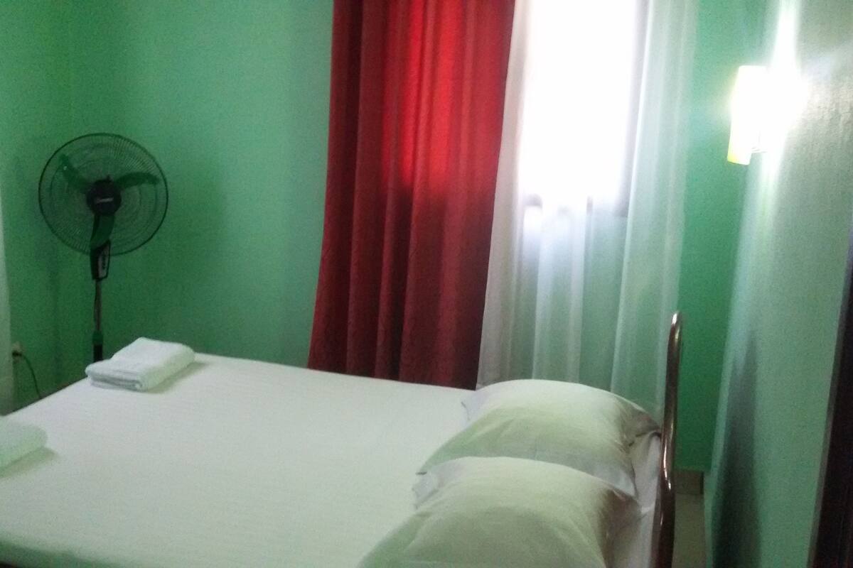 Ein kleiner, schlichter Raum mit einem Bett, einem weißen Kopfkissen, einer weißen Decke, einer grünen Wand, einem roten Vorhang und einem Standventilator.