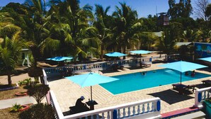 A swimming pool with blue umbrellas, surrounded by palm trees and a white fence. - Saara Hotel (Mahajanga)