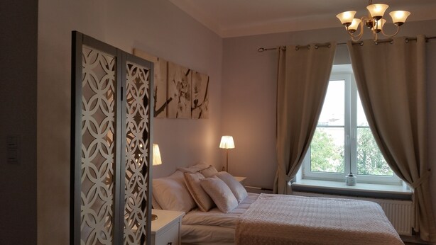 A bedroom with a bed, a nightstand, a lamp, a window with curtains, and a decorative screen. - Old Town Art Apartment (Warsaw)