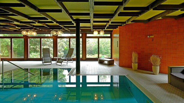 Piscina con piso de vidrio, vigas de madera en el techo y grandes ventanales que ofrecen vista a la vegetación.