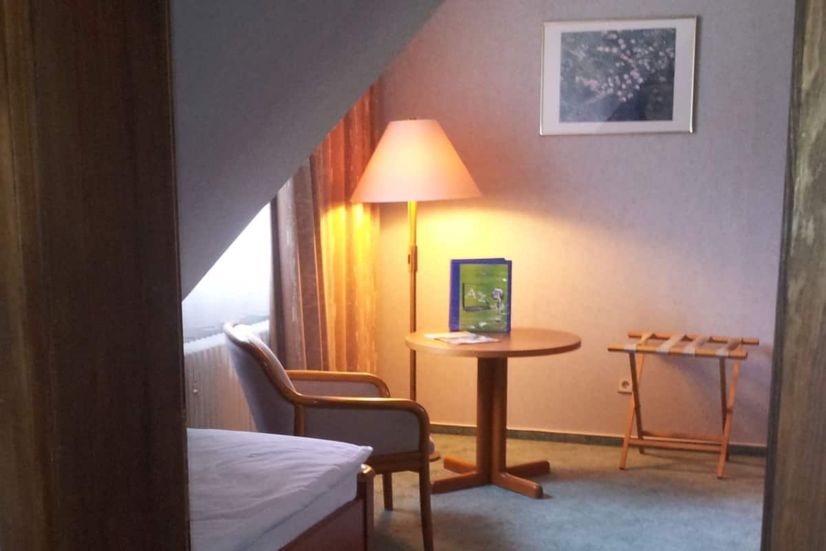 A small, dimly lit room with a single bed, a chair, a small table, and a lamp.
