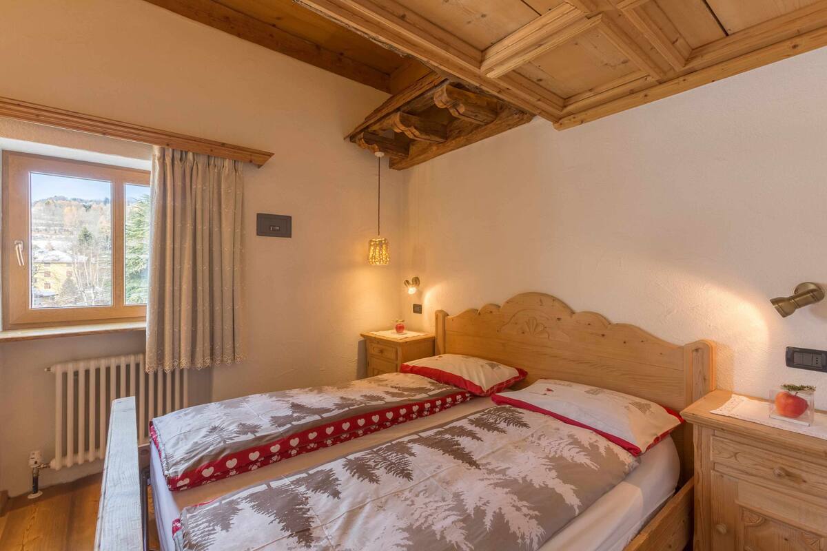 A bedroom with a wooden headboard, a bedside table with a lamp, a window with curtains, and a radiator.