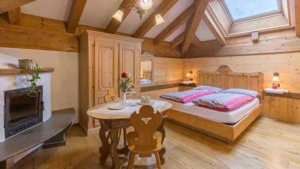 A cozy room with a wooden ceiling, a skylight, a bed with pillows, a bedside table, a small table with a vase, and a fireplace. - Historic Hotel La Stua (Cavalese)