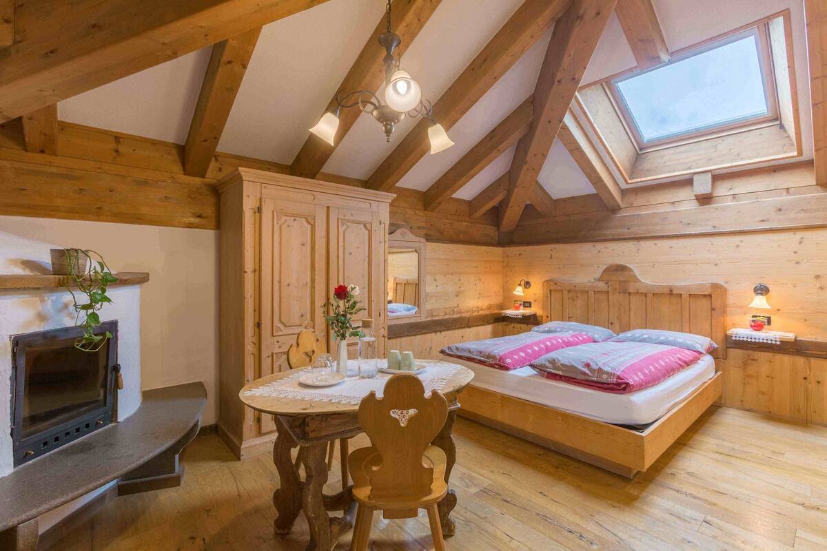 A cozy room with a wooden ceiling, a skylight, a bed with pillows, a bedside table, a small table with a vase, and a fireplace.