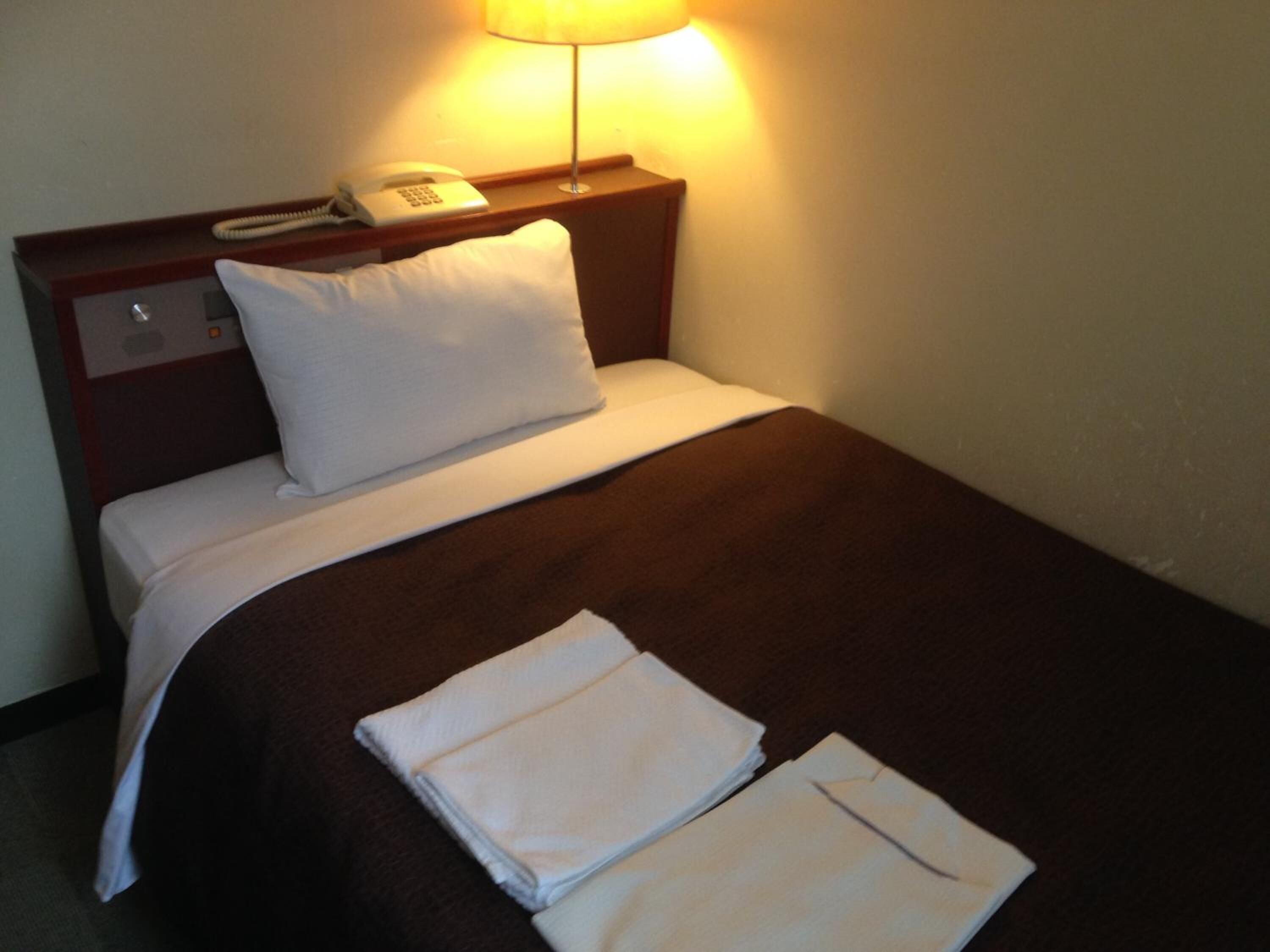Una cama bien hecha con una cubierta marrón, almohadas blancas y toallas blancas dobladas. Hay una mesita de noche con una lámpara, un teléfono y un pequeño reloj.