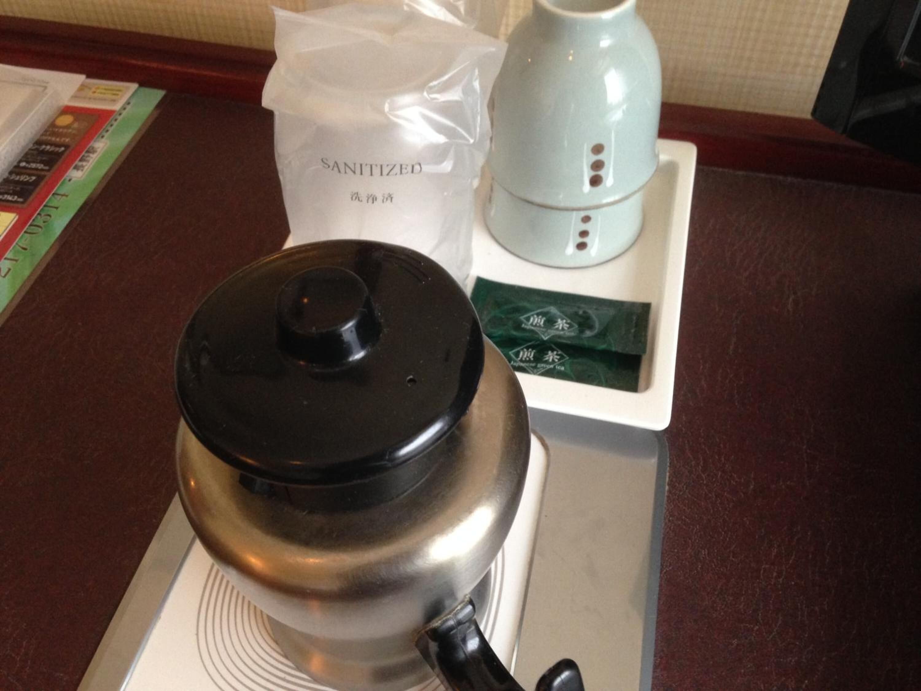 A stainless steel teapot with a black lid on a white tray, accompanied by a white bag labeled "SANITIZED" and a small white appliance with buttons.