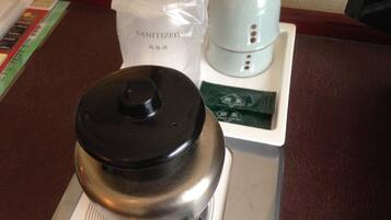 A stainless steel teapot with a black lid on a white tray, accompanied by a white bag labeled "SANITIZED" and a small white appliance with buttons.