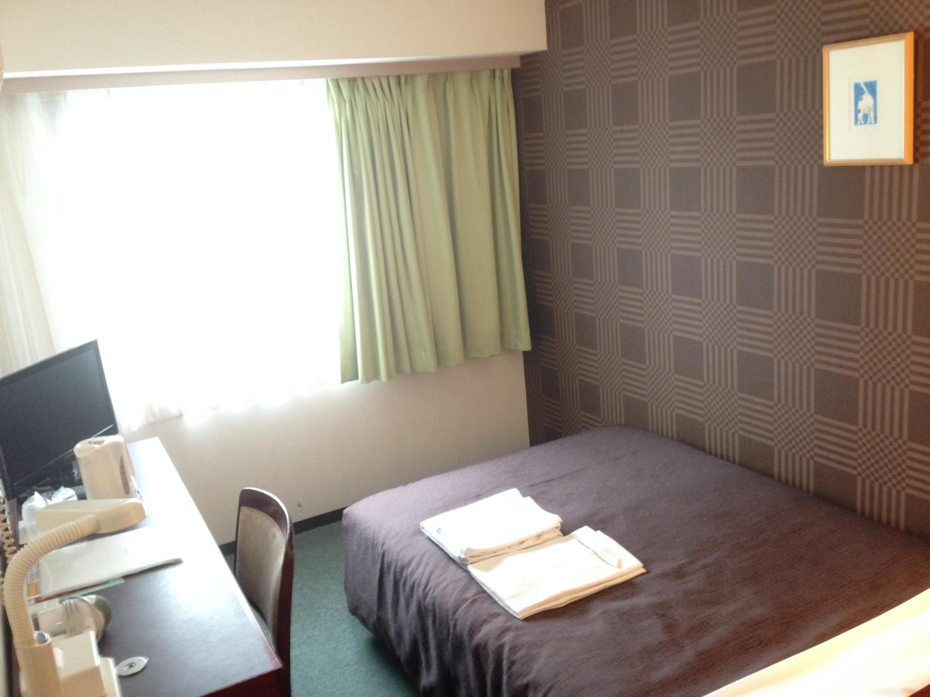 A hotel room with a bed, a desk with a telephone, a chair, a window with curtains, and a patterned wall.
