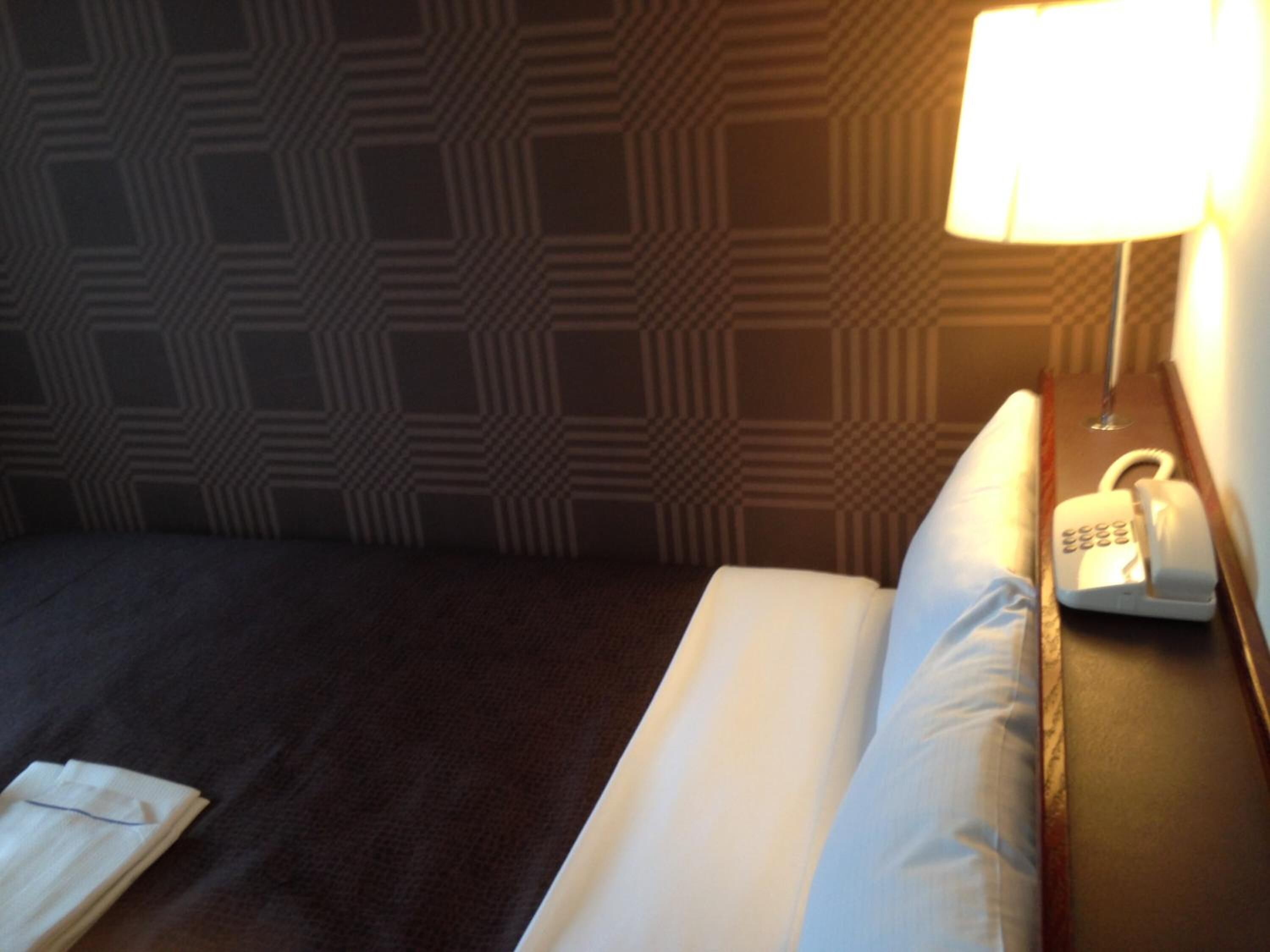 A hotel room with a neatly made bed, a bedside lamp, and a telephone on the nightstand.