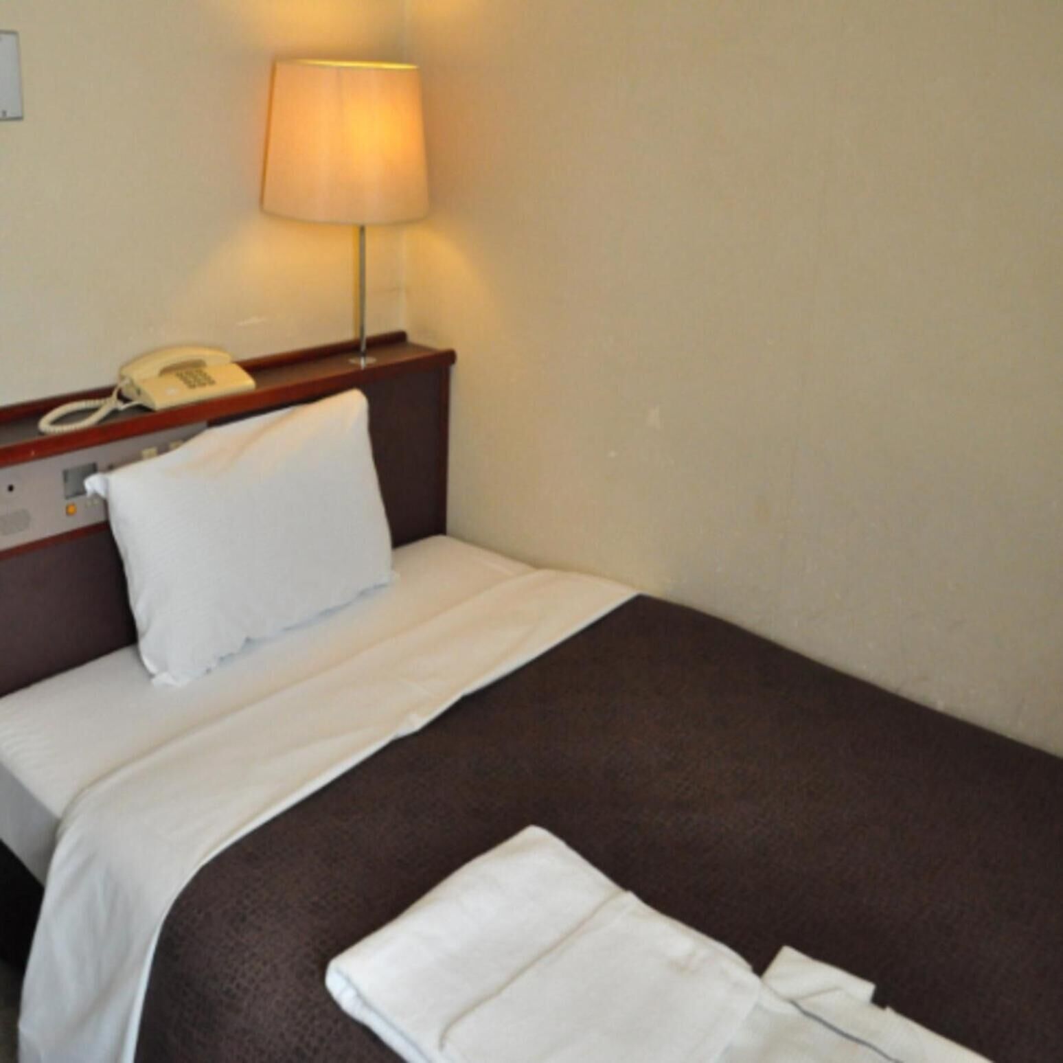 A hotel room with a bed, a bedside lamp, a telephone, and a folded towel on the floor.
