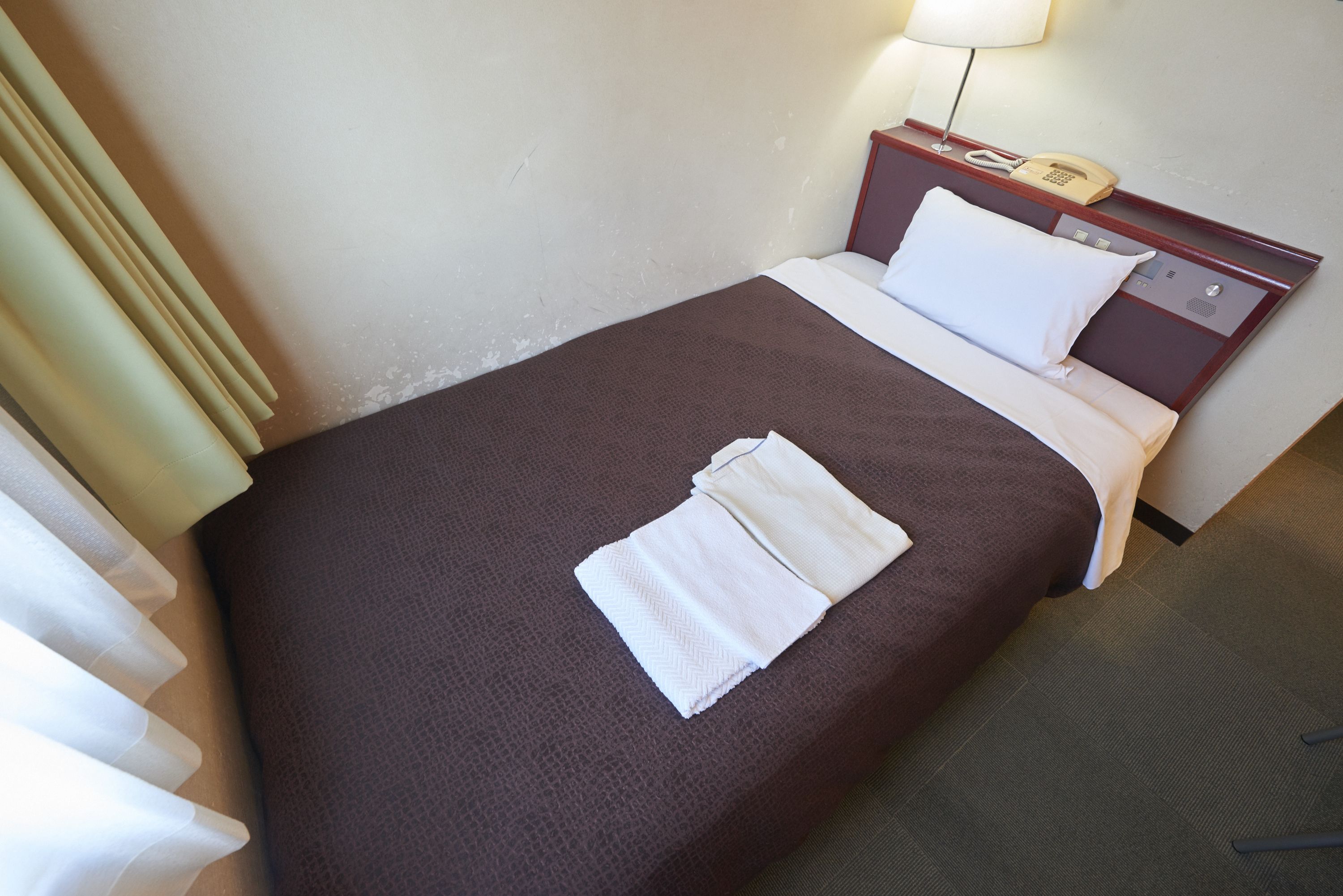 A neatly made bed with a brown bedspread and white pillows, a bedside telephone, and a lamp on a wooden nightstand.