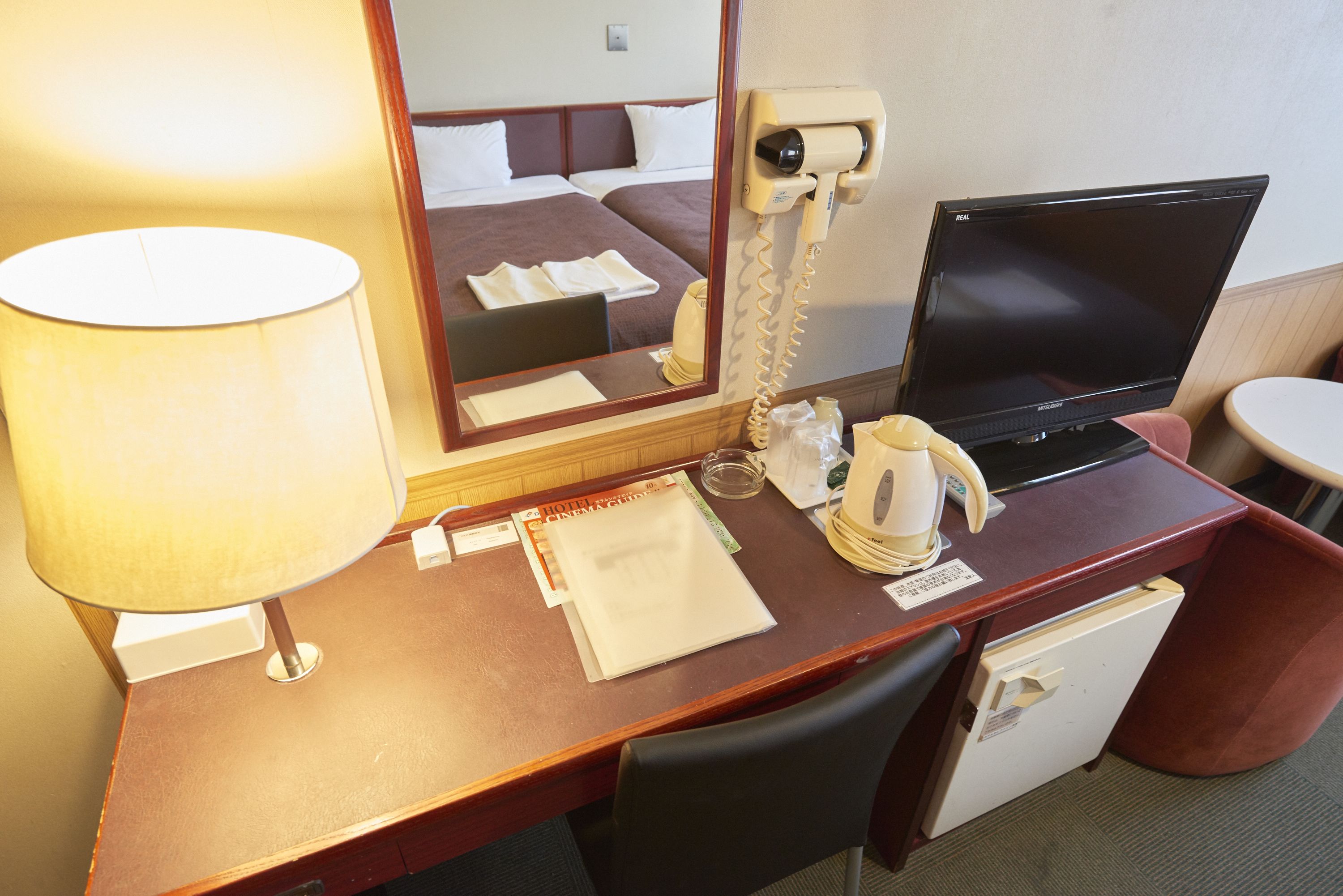 A hotel room desk with a lamp, a mirror, a laptop, a phone, a coffee maker, and a small refrigerator.