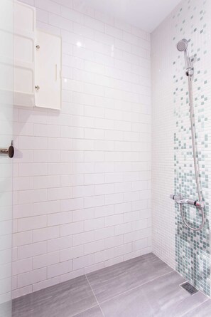 Un baño con paredes de azulejos blancos, una ducha con detalles de mosaicos y piso gris.