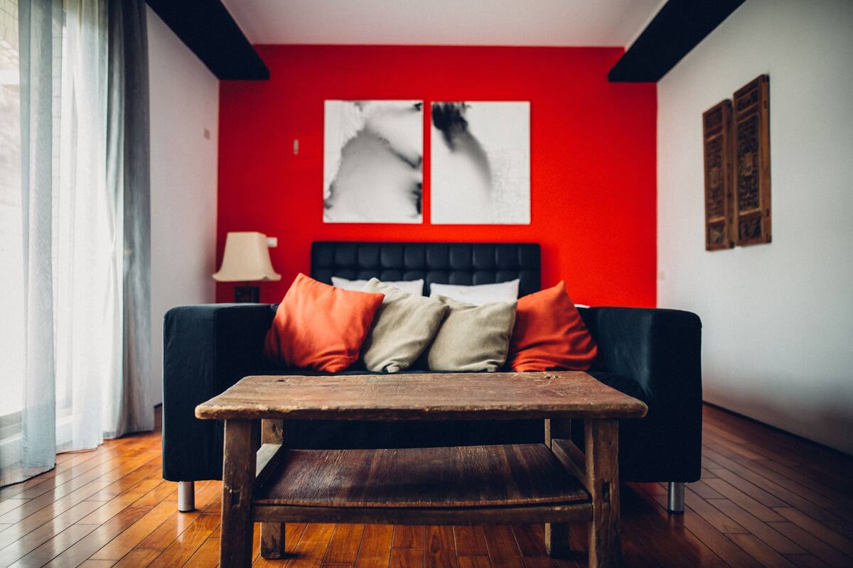 A modern room with a red accent wall, a black sofa with orange and beige cushions, a wooden table, and a lamp.