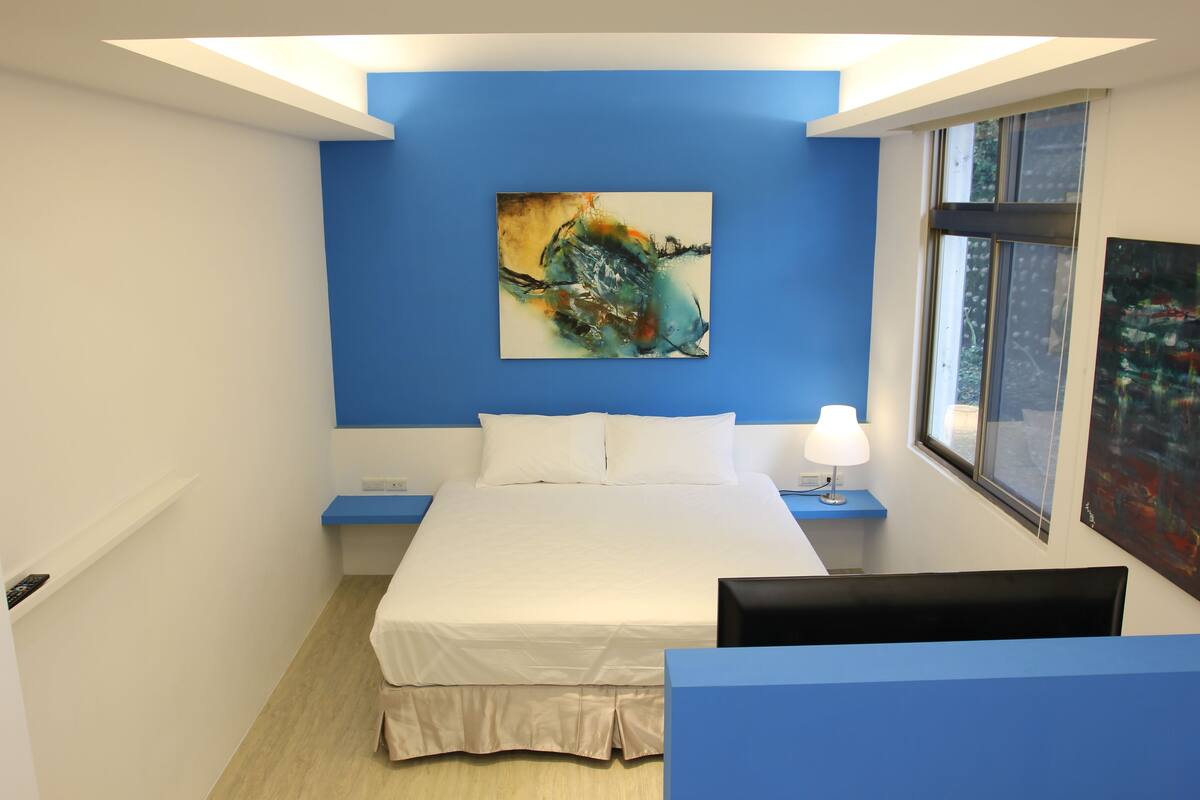A modern bedroom with a blue accent wall, a large abstract painting, a white bed, and a window with a view.