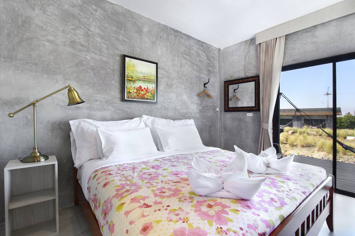 A modern bedroom with a large bed, a bedside table, a lamp, and a view of the outdoors.