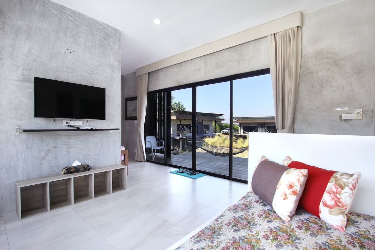 A modern living room with a flat-screen TV, a sofa with floral cushions, a bench with storage, and a large sliding glass door leading to an outdoor area.