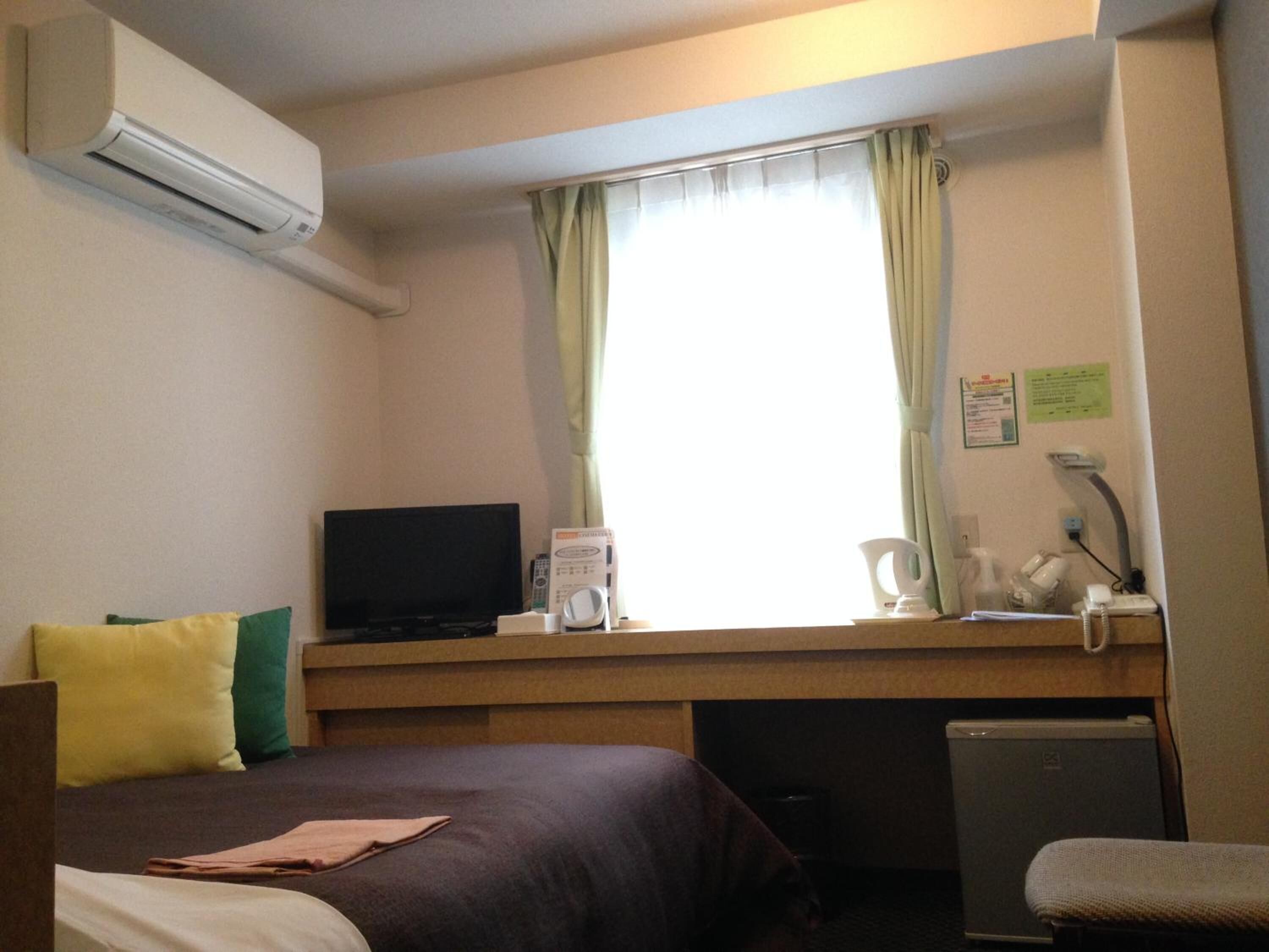 Habitación de hotel con cama, escritorio con televisor, refrigerador pequeño y ventana con cortinas.