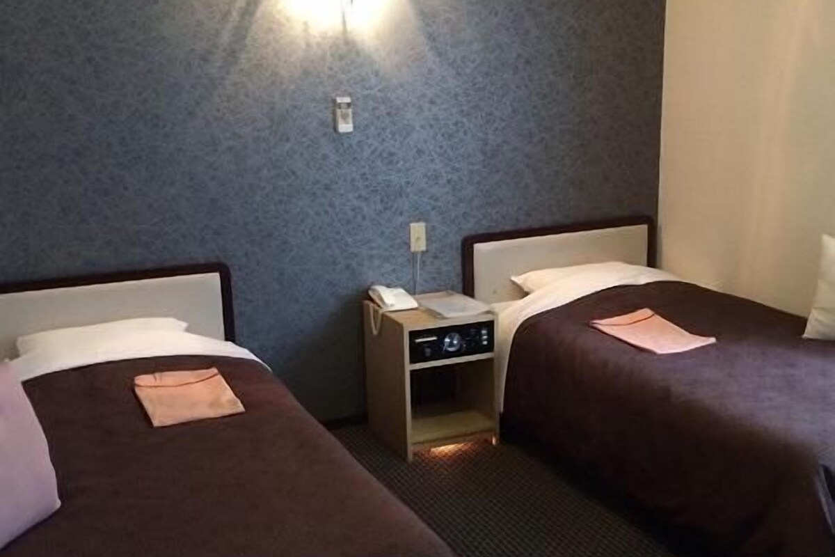 A hotel room with two beds, a small bedside table with a lamp, and a wall-mounted light fixture.