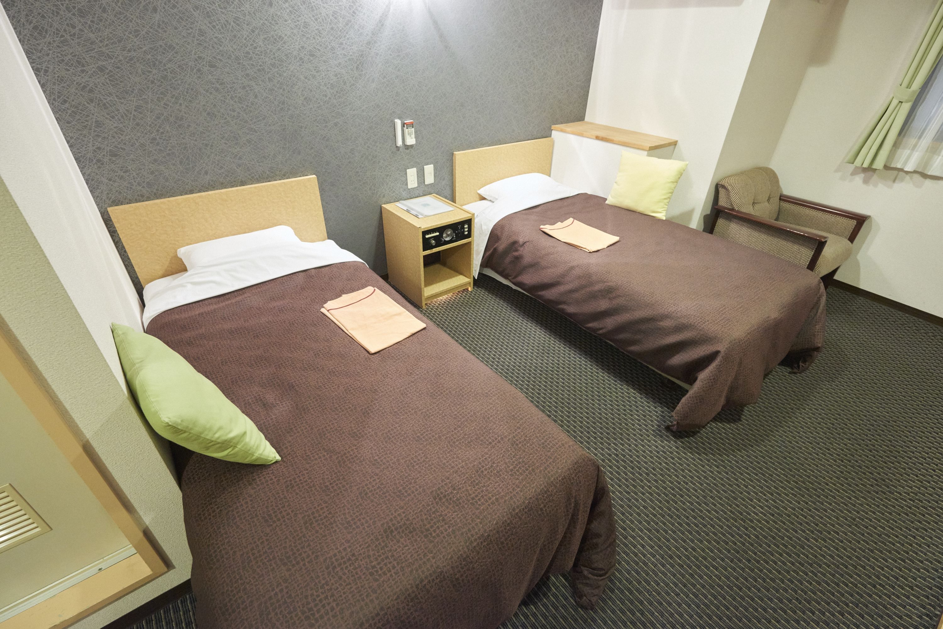 Habitación de hotel con dos camas individuales, una mesita de noche pequeña con una radio, una silla y una ventana con cortinas.