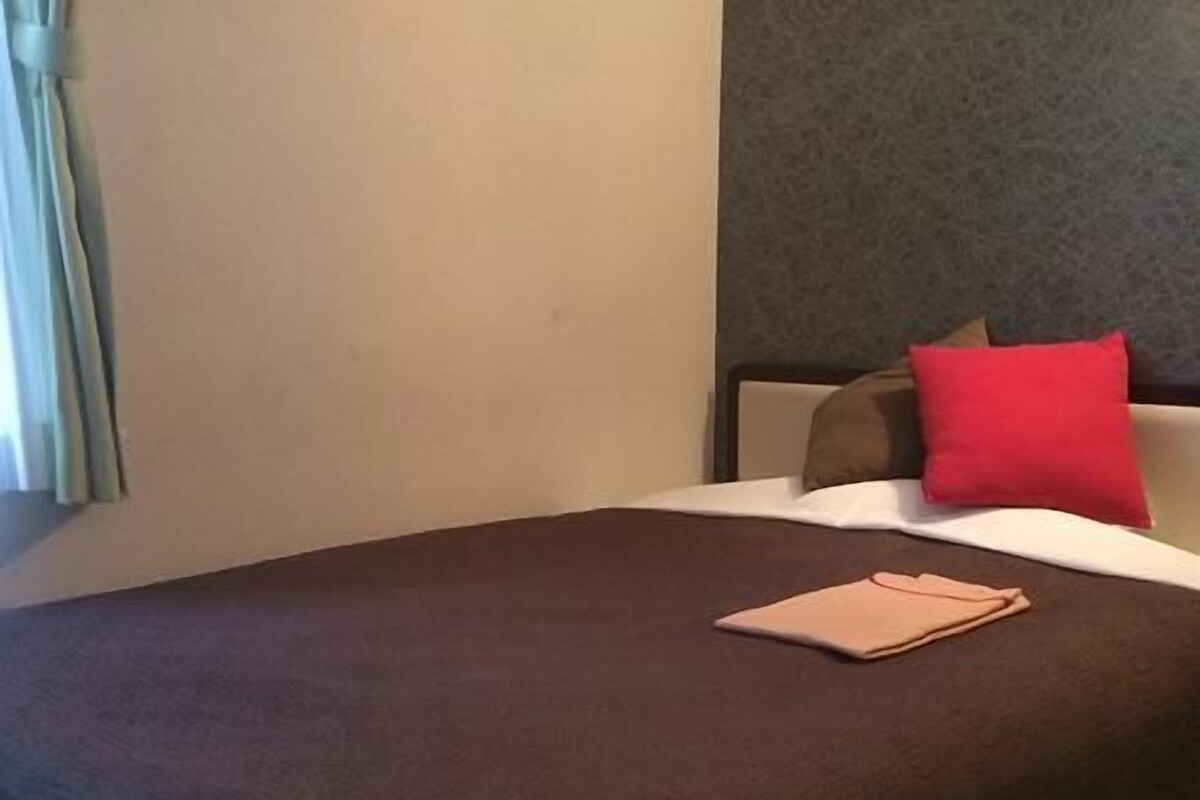 A hotel room with a large bed, a red pillow, and a beige folder on the bed.