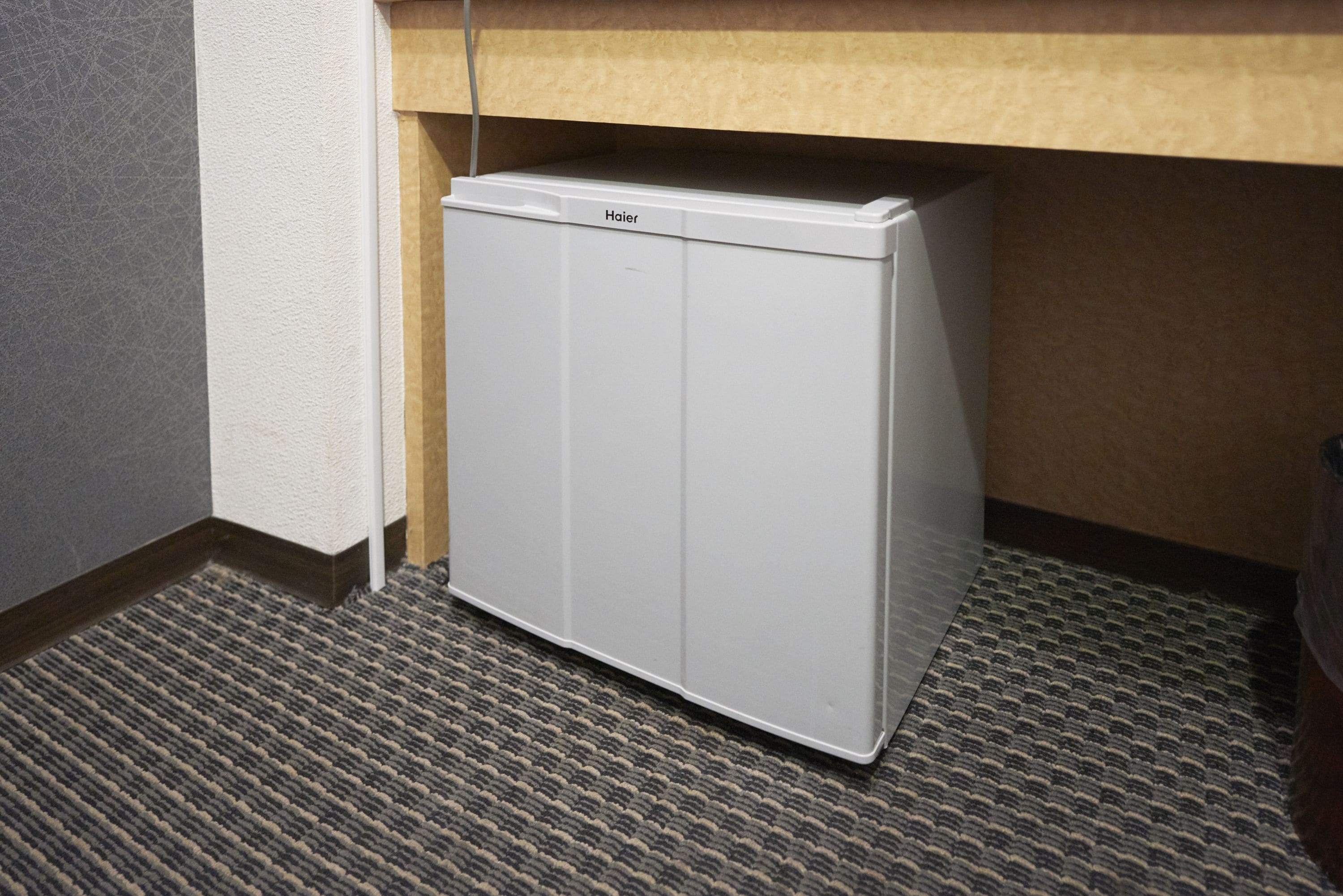 A compact white refrigerator under a wooden counter in a hotel room.