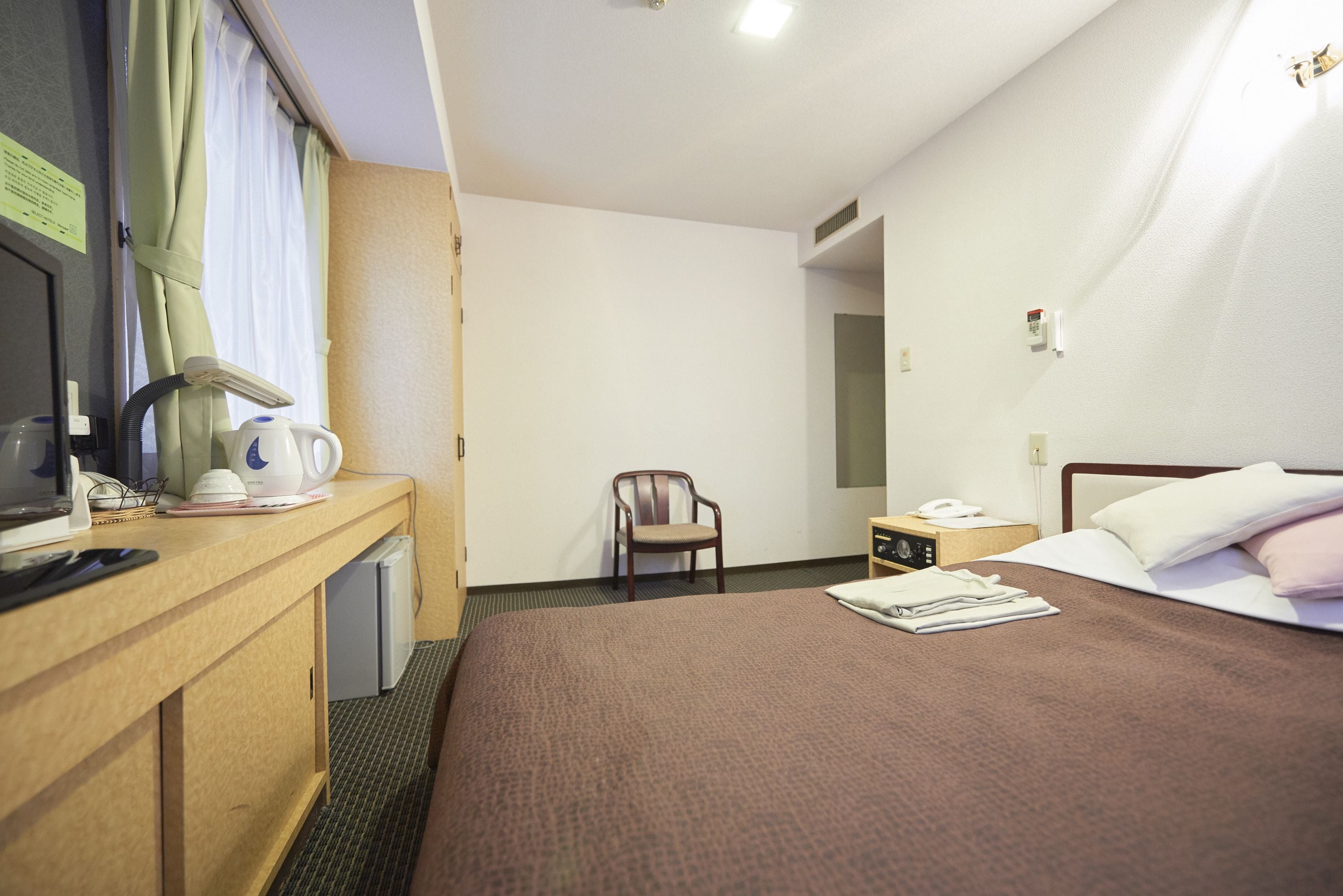 A hotel room with a bed, a desk with a computer, a chair, and a nightstand with a phone.