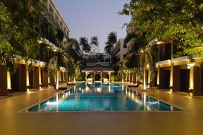 A swimming pool with a covered pavilion in the center, surrounded by palm trees and buildings with warm lighting. - Summer Tree Hotel (Bangkok)