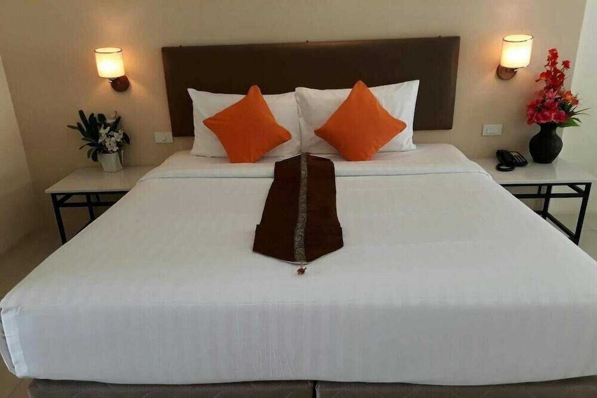 A neatly made bed with white and orange pillows, a brown throw, and a headboard. There are two bedside tables with lamps and a vase of flowers.