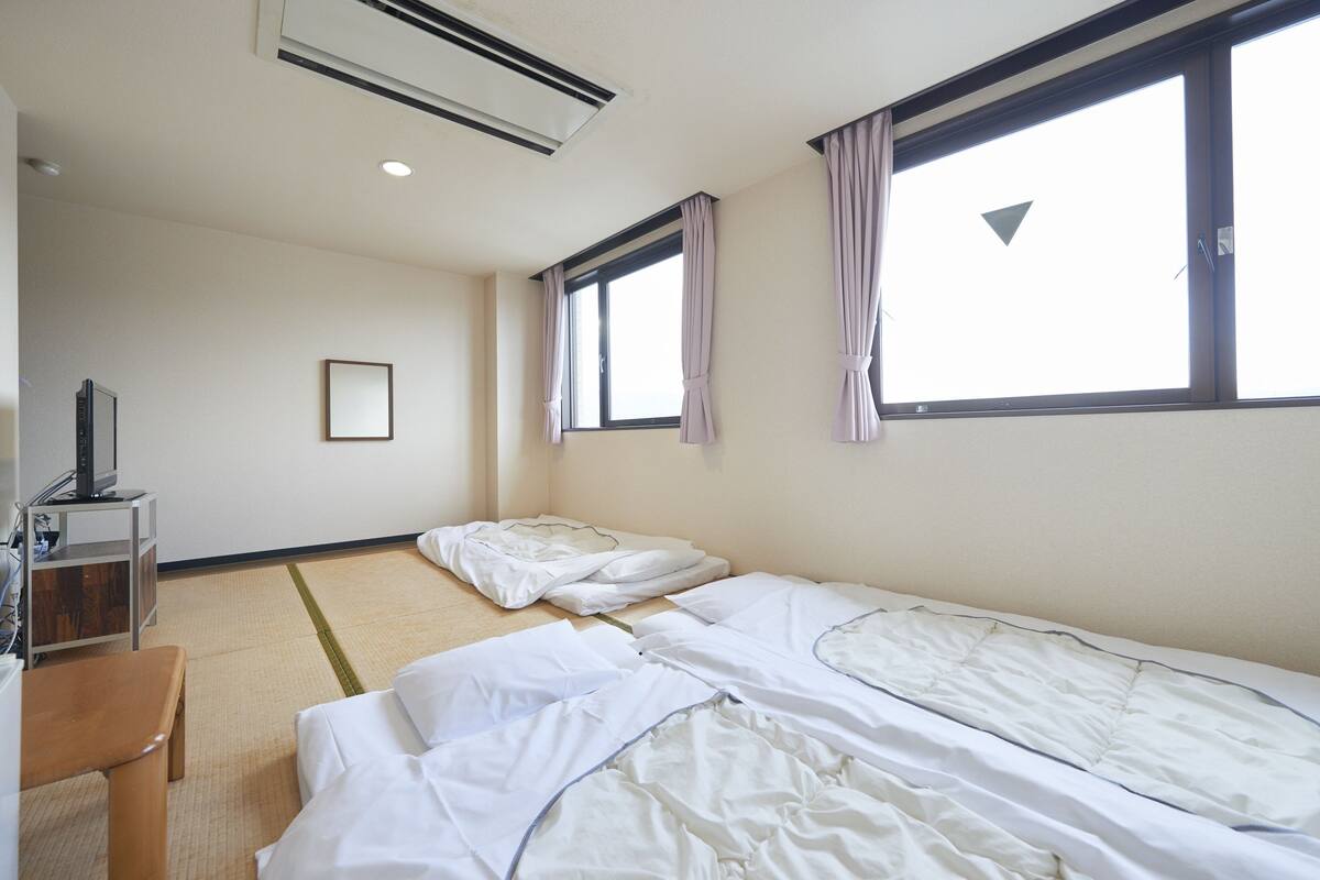 Habitación de hotel con dos camas individuales, un banco de madera, una mesita con televisor y una ventana con cortinas.