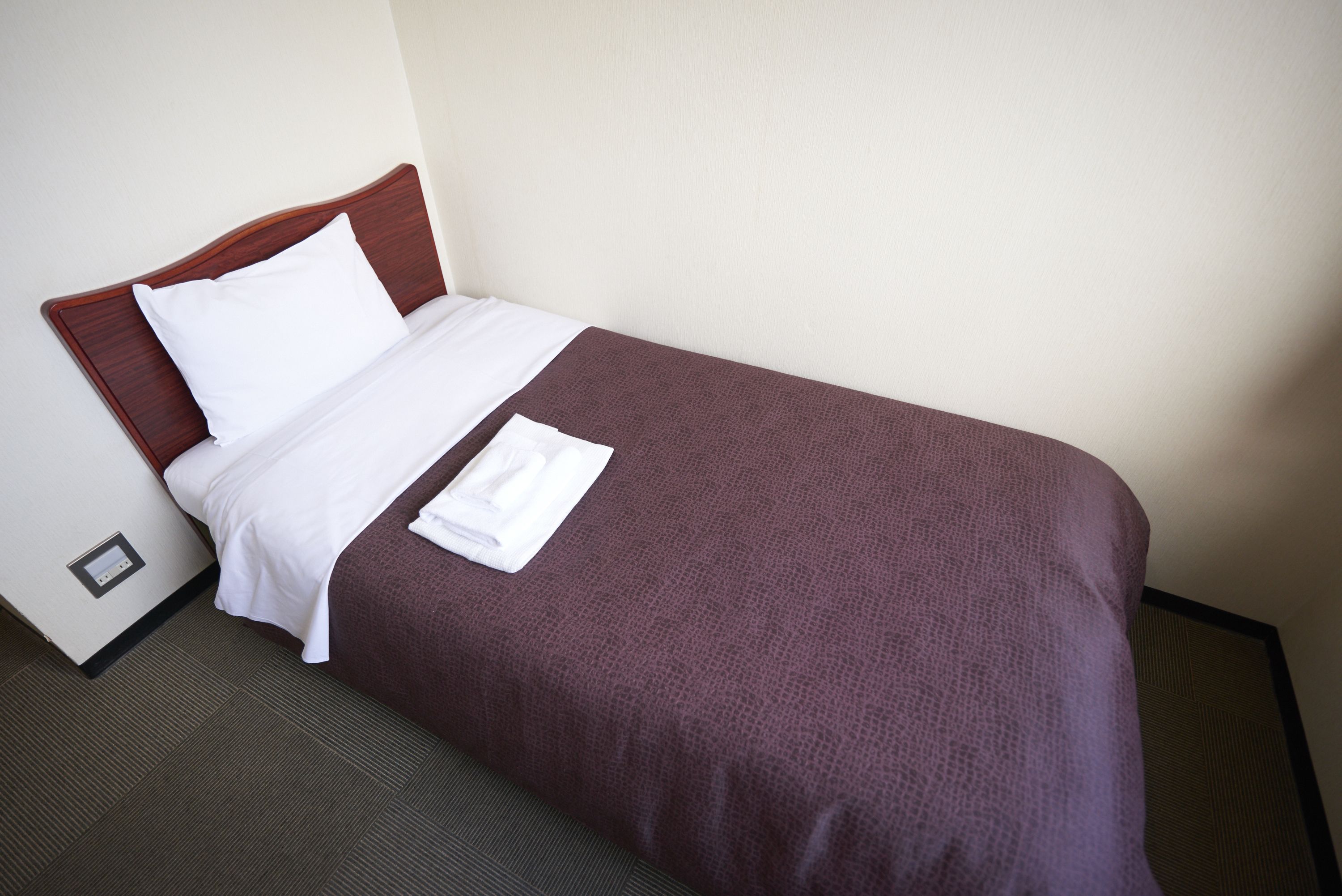 A neatly made bed with a brown cover and white linens, a power outlet on the wall, and a dark carpeted floor.