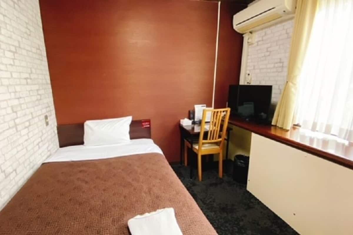 A hotel room with a single bed, a desk with a chair, a television, and an air conditioning unit.